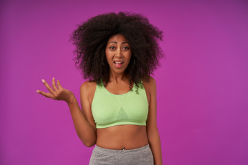 Puzzled young curly female with dark skin wearing casual hairstyle, raising palm perplexedly and looking to camera with raised eyebrows, isolated over purple background