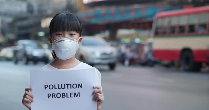 Little girl in protective mask holding pollution problem sign at city.