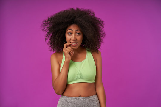 Pretty Young Dark Skinned Woman With Curly Hair Wearing Casual Sporty Clothes, Looking To Camera With Raised Eyebrows And Keeping Forefinger On Underlip, Standing Over Purple Background