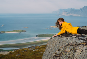 Smiling girl in yellow jacket and sunglasses lies on stone in on the edge of rocks in mountains and looking to fjord