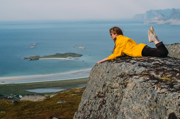 Girl in yellow jacket and sunglasses lies on stone in on the edge of rocks in mountains and looking to fjord