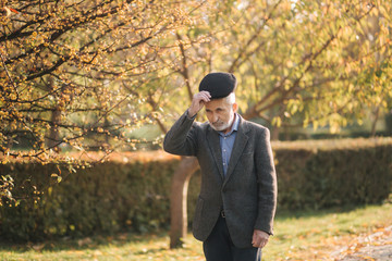 Handsome elderly man putting on a gray jacket and black hat. Old gray-haired bearded man walk in the autumn park. Yellow background