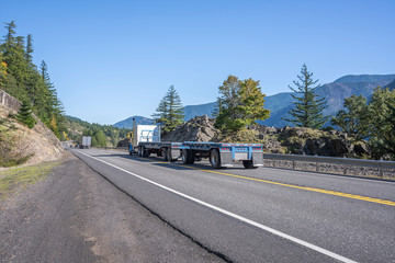 Big rig blue classic semi truck transporting two empty flat bed semi trailers going to warehouse for load running on the road along the Columbia Gorge area © vit