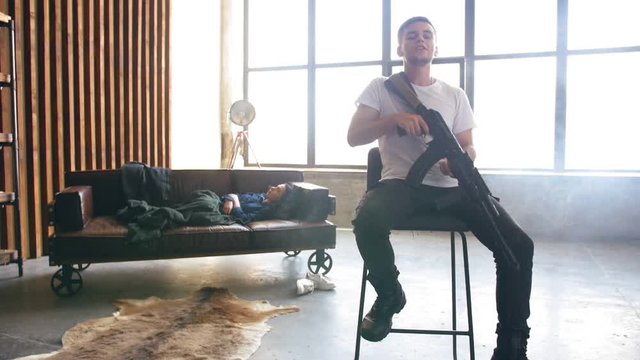 Young man sits on a chair holding a machine gun in his hand and read rap. The girl getting up from the couch in the background in defocus. Full shot.