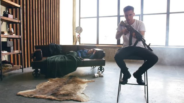 Young man sits on a chair holding a machine gun in his hand and read rap. The girl getting up from the couch in the background in defocus. Full shot.