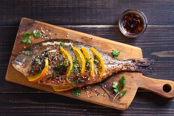 Orange ginger sea bream fish, sprinkled with sesame seeds and cilantro on serving board. Soy orange sauce. Overhead horizontal image