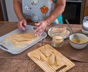 Obraz na płótnie Canvas Elderly woman hands preparing breaded fish filets with eggs. Freshness on the table with wooden chopping board and salt