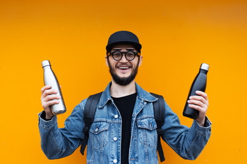 Portrait of cheerful young hipster, holding black and silver stainless thermo eco bottle for water, on yellow or orange background.
