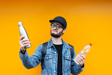 Portrait of cheerful young hipster, holding steel thermo eco bottle for water and plastic bottle,...