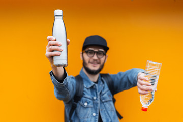 Portrait of cheerful young hipster, holding steel thermo eco bottle for water and plastic bottle,...