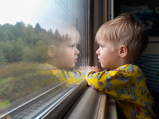 A bright, cute three-year-old boy riding a train looks out the window, behind which a dull...