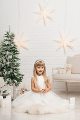 A little girl sits and holds a candle in her hand in a room near the Christmas tree.