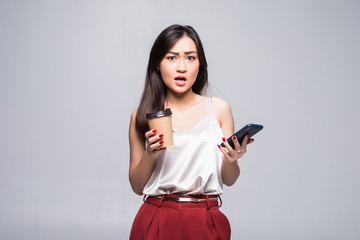 Naklejka premium Portrait of a smiling asian woman using mobile phone while holding cup of coffee to go isolated over white background