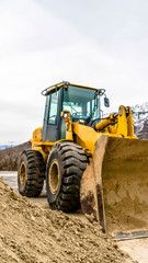 Vertical Front view of a yellow bulldozer against snow topped mountain and cloudy sky