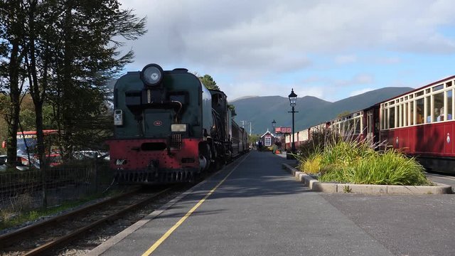 Two old steam trains at railway station platform in Wales, UK. One vehicle is standing still while another train is moving toward the camera POV. People, unrecognizable, seen in a distance and inside 