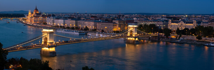 Fototapeta premium Panorama of Budapest along the Danube River featuring the Chain Bridge and Hungarian Parliament Building at blue hour. 