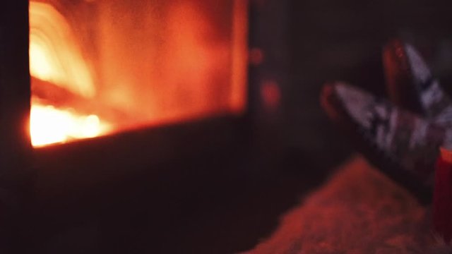 Feet in woollen socks by Burning Christmas Fireplace. 4K STABILIZED shot. Woman relaxes by warm fire with a cup of hot drink and warming up her feet. Close up. Winter and Christmas holidays concept.