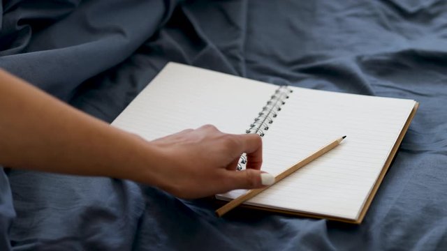 hand placing pencil on diary notebook on blue bedding, concept of journaling