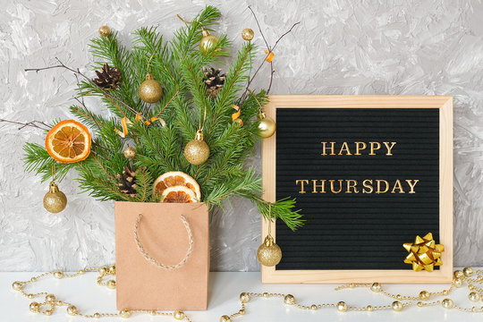 Happy Thursday text on black letter board and festive bouquet of fir branches with christmas decor in craft package on table. Template for postcard, greeting card Concept Hello winter Thursday