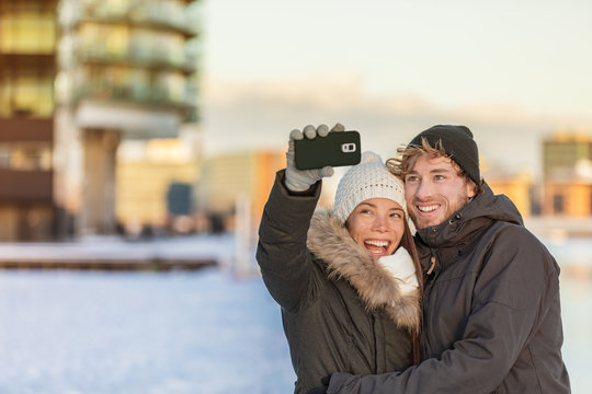 Selfie couple happy winter travel walk tourists taking photo with phone on city street panorama lifestyle. Asian woman, Caucasian man wearing knit hats and jackets. - Powered by Adobe
