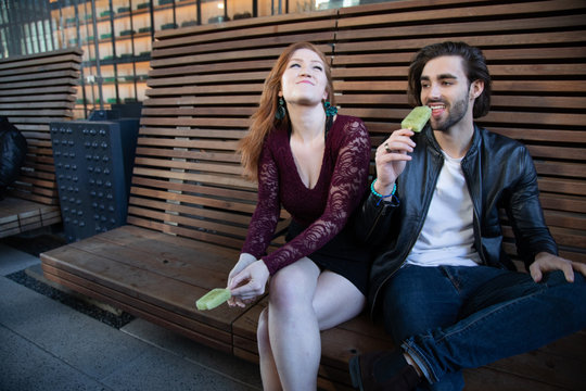 Young Couple Sitting On Bench Laughing And Eating Green Popsicles