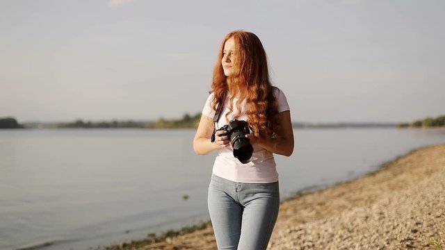 Sun-kissed freckled redhead girl with photo-camera is walking on bank of river.