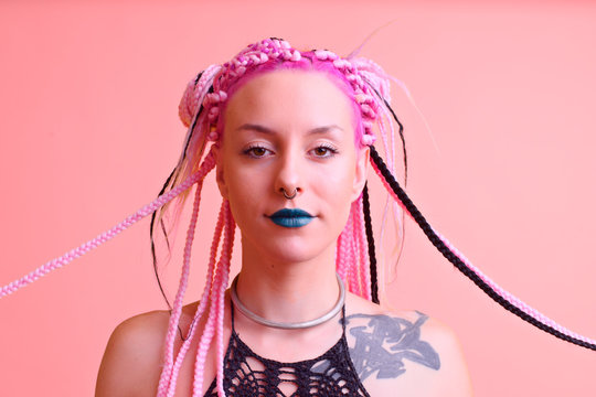 portrait of a modern and tattooed young girl in alternative style with colored braids posing in the studio in front of a pink background