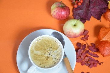 White cup of coffee with cappuccino cream on an orange background with autumn leaves, apples and red berries. Fall season, leisure time and coffee break concept.