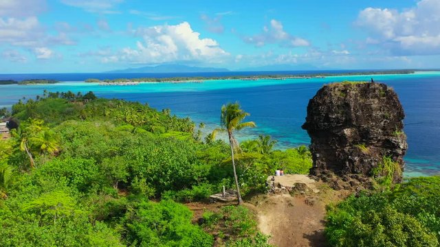 Aerial shot from the back of a young couple sitting next to a ruin wall on a beautiful tropical island, drone ascending the flying backwards - Bora Bora, French Polynesia
