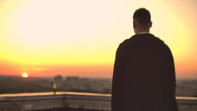 Back view a male freelance programmer in a hoodie walks on the roof at sunset and drinks beer looking at the beautiful view of the city