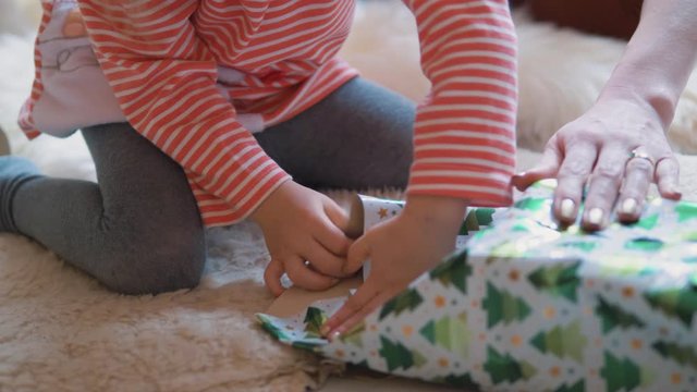 cute little girl wrapping christmas or birthday gift with her mother. Family sitting on a white carpet preparing presents for friends and family. Close up from hands.