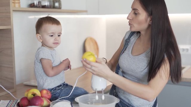 Cute little boy toddler and his mother is cooking in the modern kitchen together. Mother is mixing a dough in a bowl with a mixer and son is trying to bite an apple.