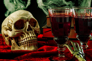 Halloween backdrop, haunted house and vampire drink conceptual idea with skull, glass filled with blood, spiderwebs, vintage books and skeleton arm on black background