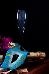 CARNIVAL MASK NEXT TO CHAMPAGNE BOTTLE, GLASS CUP AND TREE LEAVES. CARNIVAL PARTY