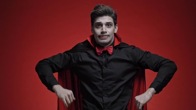 Vampire man with blood and fangs in black halloween costume touching his head and screaming because of getting bad news isolated over red wall