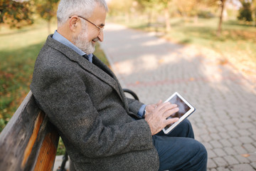 Fototapeta premium Handsome gray-haired elderly man with beautiful beard sitting on the bench and use tablet for scrolling in internet. Background of autumn tree