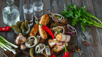 Vodka with lard, salted fish and vegetables, sausages on wooden background. Alcohol pure craft drink and traditional snack, tomatos, onion, cucumbers. Negative space. Celebrating food and delicious.