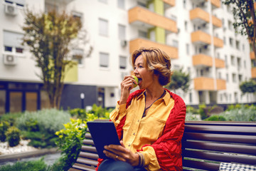 Charming caucasian fashionable senior woman with blonde short hair sitting on bench in park, eating apple and using tablet.