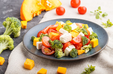 Vegetarian salad with broccoli, tomatoes, feta cheese, and pumpkin on a blue ceramic plate on a black concrete background, side view, close up, selective focus.