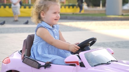 little girl in a blue dress rides a pink baby car in a park. Happy childhood concept. Caricature of an adult woman driving a car.