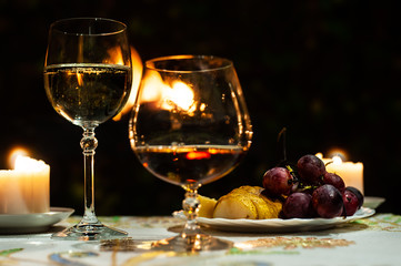 Two glasses with white sparkling wine and brandy or whiskey on table with fruits plate and big fire on the black background