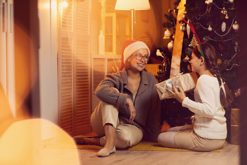 Girl giving Christmas gift box to her happy mother and congratulating her with Christmas while they sitting on the floor near the Christmas tree