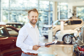 A car dealer smiling standing in a showroom. - obrazy, fototapety, plakaty