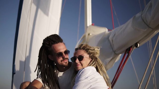 Low angle footage of stylish couple with dreadlocks in white clothes and sunglasses sitting embracing on the bow of the yacht and kissing. Loving couple spending their honeymoon on the yacht beyond