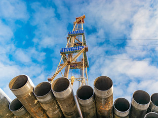 In the foreground pipes for drilling oil and gas wells. In the background, a blurred image of the rig out of focus. Blue sky with clouds.