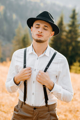 Pretty groom in white shirt holds suspenders with hands on mountain background. Rustic style