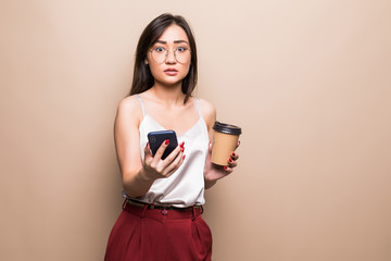 Full length portrait of a smiling asian woman using mobile phone while holding cup of coffee to go isolated over beige background