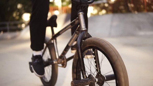 Unrecognizable extreme bmx biker pedals his black bike in a sunny skate park in the summer. Cool young bmx biker cycling outdoors. Close up of the wheels. Slow motion