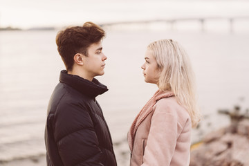 Fototapeta premium Lovely brunette guy and pretty girl blonde look at each other in eyes, in love look. Loving teenagers are happy, smiling, kissing. Teen Love Concept. Outdoor.