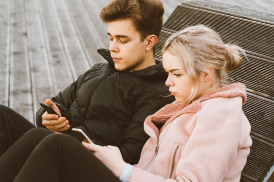 Loving teenagers on date look at mobile phones, sit on park bench in fall. Immersion in virtual world, social networks. Concept of teenage love, Smombie. Teen dating.